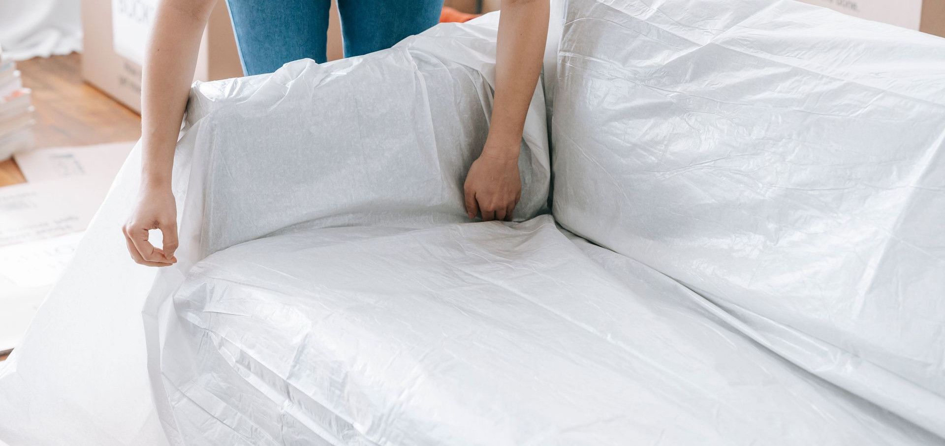 A woman carefully wraps a sofa in protective material while packing boxes in a bright room.